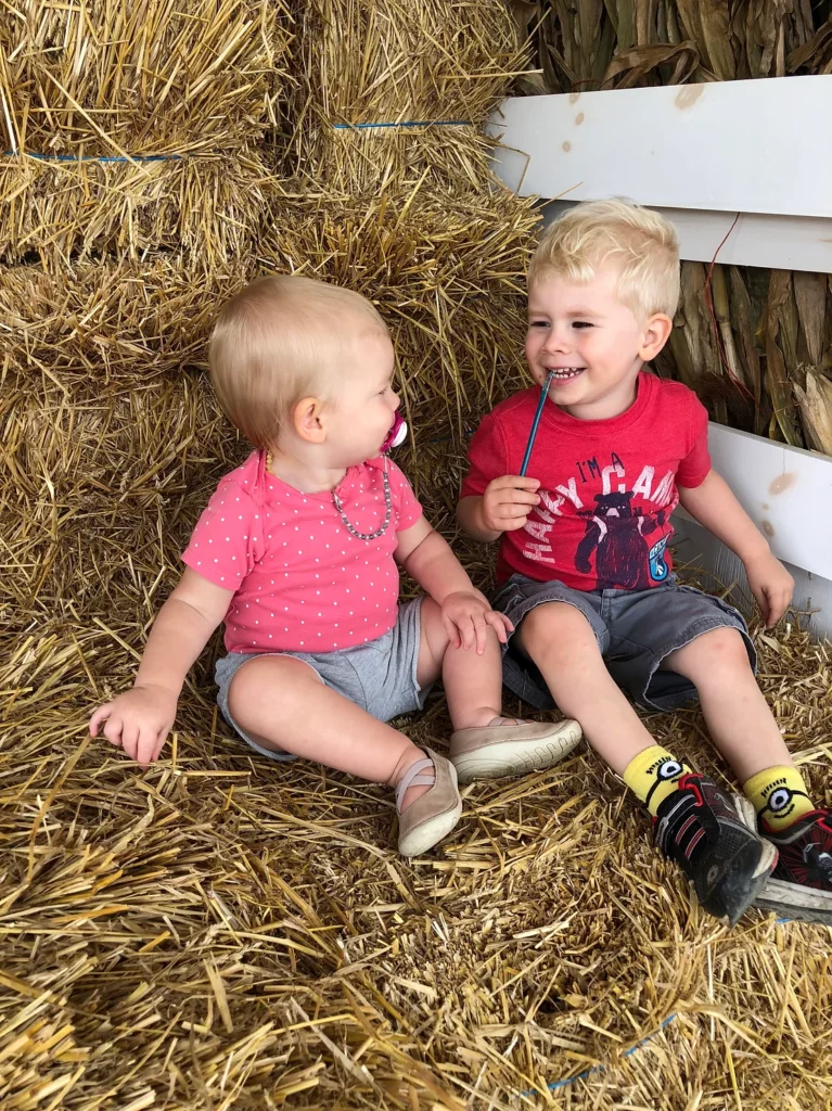 brother and sister sitting on hay bale. brother is smiling at sister.